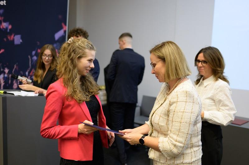 zdjęcie sali, w której odbywa się konferecja. Widać gości i gościnie siedzących przy stołach, a tle siedzą prowadzace spotkanie