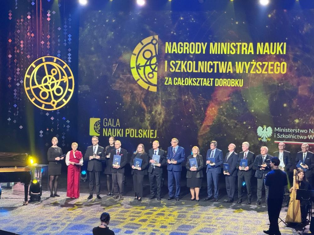 Kilkanaście elegancko ubranych osób stoi na scenie, trzymając nagrody. W tle napis „Nagrdy Ministra Nauki i Szkolnictwa Wyższego za całokształt dorobku. Gala Nauki Polskiej